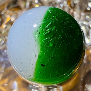 May include: A glass marble with a white and green design. The green side has a textured surface with small bubbles.