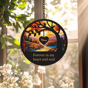 May include: A wooden circle with a stained glass design of a tree and a sunset. The circle is hanging from a chain and has the words "Forever in my heart and soul" written on it. There is a small heart with the word "Mom" on it hanging from the tree.