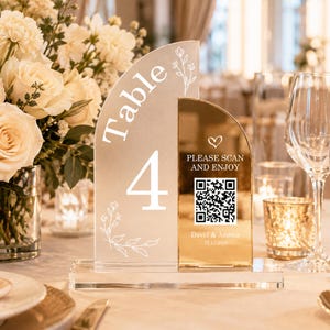 May include: A dual-panel acrylic table number sign featuring a frosted arch with the text Table 4 and floral line art, paired with a gold mirrored arch displaying a QR code, the text PLEASE SCAN AND ENJOY, David & Amelia, and 12.12.2026.
