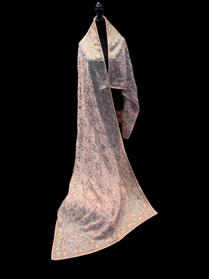 Op de afbeelding: Een lichtroze sjaal met ingewikkelde bloemenborduurwerk en een delicate rand. De sjaal is over een zwarte mannequin gedrapeerd, wat het vloeiende ontwerp en het gedetailleerde vakmanschap benadrukt. De borduursels bevatten zilveren en gouden draden.