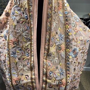 Op de afbeelding: Lichtroze, openvallend kledingstuk met ingewikkelde borduursels. De borduursels tonen bloemen- en paisley-motieven in blauw, geel en roze, geaccentueerd met gouden randen en kleine spiegelversieringen. Het kledingstuk heeft een satijnachtige afwerking.