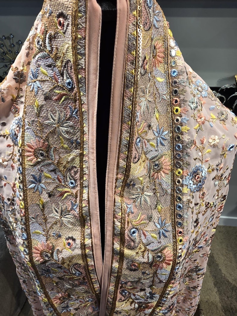 Op de afbeelding: Lichtroze, openvallend kledingstuk met ingewikkelde borduursels. De borduursels tonen bloemen- en paisley-motieven in blauw, geel en roze, geaccentueerd met gouden randen en kleine spiegelversieringen. Het kledingstuk heeft een satijnachtige afwerking.