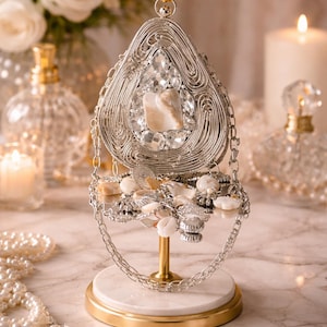 May include: A silver jewellery display stand with a gold base and a white marble accent. The stand holds various necklaces, rings, and earrings. The display features a large, teardrop-shaped wireframe design with a central stone and surrounding crystals.