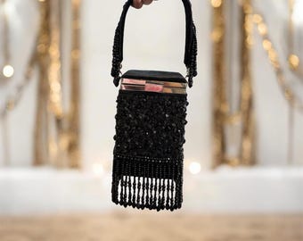Puede incluir: Un bolso de mano negro con cuentas con un cuerpo rectangular y flecos de cuentas. El bolso tiene un asa y un cierre metálico. El bolso está cubierto de pequeñas cuentas negras brillantes.