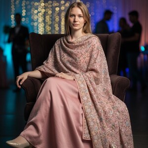 Op de afbeelding: Een vrouw draagt een lichtroze jurk en een bijpassende sjaal met ingewikkelde borduursels en versieringen. De sjaal is over de schouders gedrapeerd. Ze zit in een bruine fauteuil.