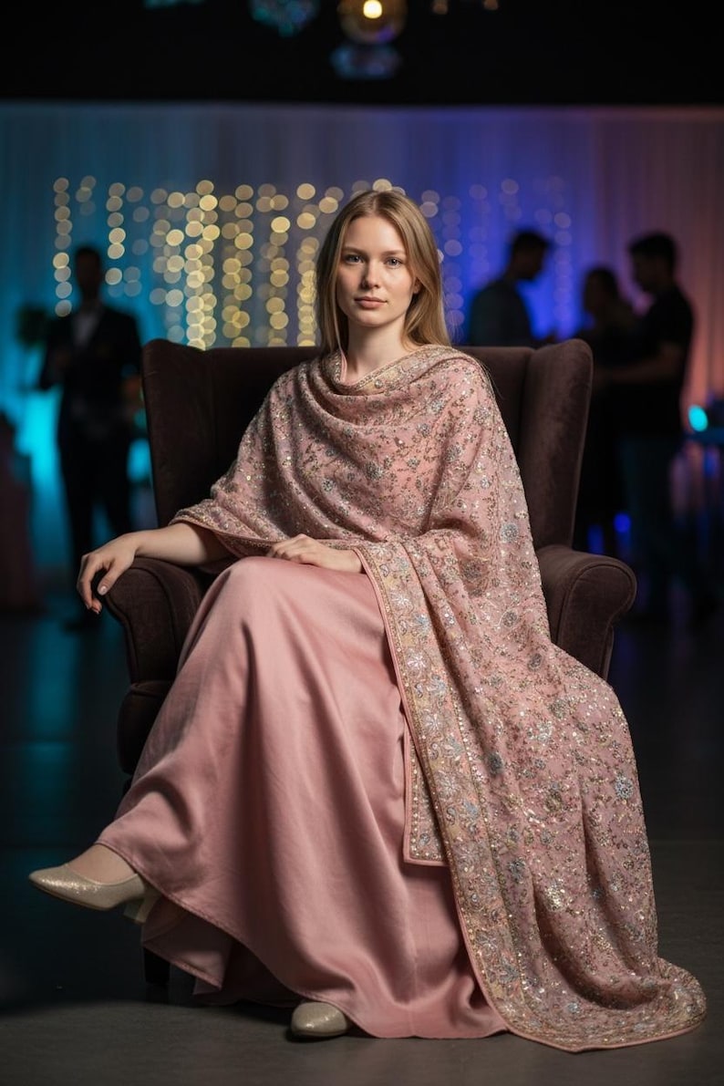 Op de afbeelding: Een vrouw draagt een lichtroze jurk en een bijpassende sjaal met ingewikkelde borduursels en versieringen. De sjaal is over de schouders gedrapeerd. Ze zit in een bruine fauteuil.