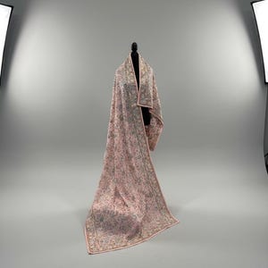 Op de afbeelding: Een lange, roze sjaal met ingewikkelde bloemenborduurwerk en een delicate rand. De sjaal is over een zwarte mannequin gedrapeerd, wat het vloeiende ontwerp en het vakmanschap benadrukt. Het kleurenpalet omvat roze, groen en goud.