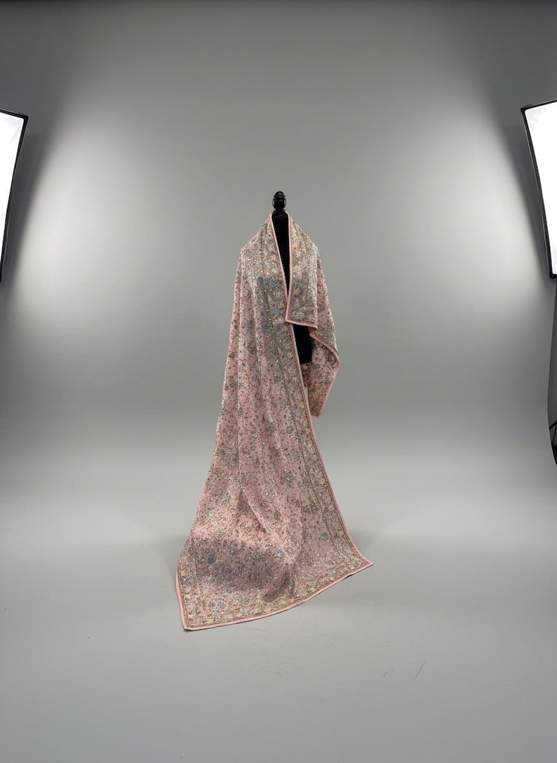 Op de afbeelding: Een lange, roze sjaal met ingewikkelde bloemenborduurwerk en een delicate rand. De sjaal is over een zwarte mannequin gedrapeerd, wat het vloeiende ontwerp en het vakmanschap benadrukt. Het kleurenpalet omvat roze, groen en goud.