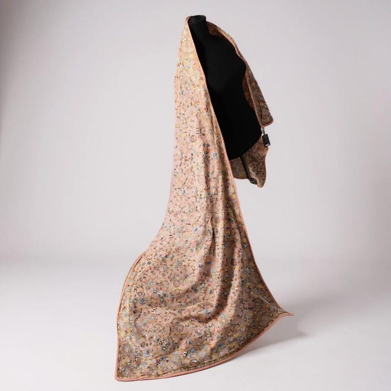 Op de afbeelding: Een lange, vloeiende sjaal met een delicaat bloemmotief in roze, blauw en geel. De sjaal is over een zwarte mannequin gedrapeerd, wat de lengte en het ontwerp benadrukt. De randen zijn afgewerkt met een subtiele rand.