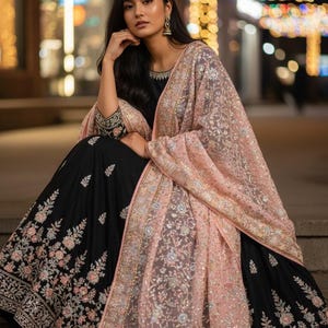 Op de afbeelding: Een vrouw draagt een zwarte jurk met bloemenborduurwerk en een lichtroze sjaal. De sjaal is versierd met ingewikkelde patronen en gouden accenten. De jurk heeft lange mouwen met gouden borduursel op de manchetten.