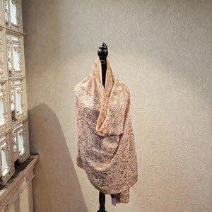 Op de afbeelding: Een perzikkleurige sjaal met ingewikkelde borduursels en versieringen is over een zwarte mannequin gedrapeerd. De sjaal heeft een gedetailleerd bloemmotief met gouden en zilveren accenten. De stof lijkt delicaat en luxueus, wat duidt op een formeel of speciaal gelegenheidskledingstuk.