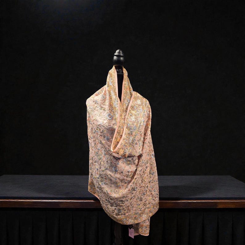Op de afbeelding: Een perzikkleurige sjaal met ingewikkelde borduursels en een delicaat bloemmotief. De sjaal is over een mannequin gedrapeerd en toont het vloeiende ontwerp en het gedetailleerde vakmanschap. De borduursels bevatten zilveren en gouden draden.