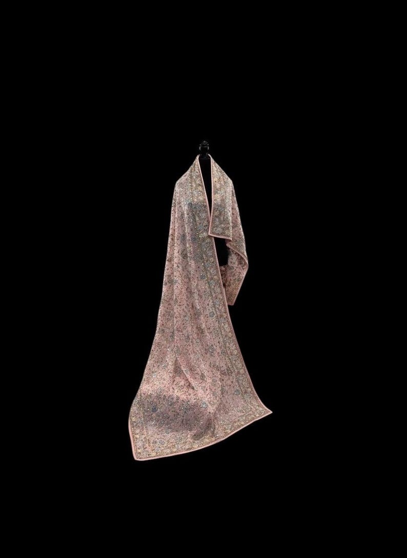 Op de afbeelding: Een lange, roze sjaal met een ingewikkeld bloemenpatroon en een contrasterende roze rand. De sjaal is over een zwarte mannequin gedrapeerd, wat de lengte en het ontwerp laat zien. De stof lijkt zacht en vloeiend.