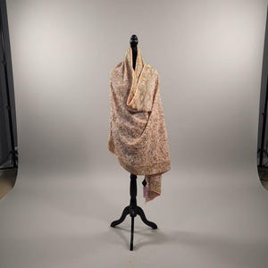 Op de afbeelding: Een lichtroze sjaal met ingewikkelde borduursels, gepresenteerd op een zwarte mannequin. De sjaal heeft een gedetailleerd bloemmotief in verschillende kleuren, wat hem een delicate en elegante uitstraling geeft. De stof drapeert sierlijk en toont zijn textuur en ontwerp.