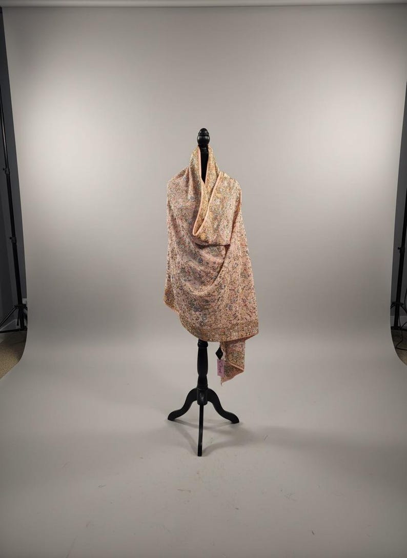 Op de afbeelding: Een lichtroze sjaal met ingewikkelde borduursels, gepresenteerd op een zwarte mannequin. De sjaal heeft een gedetailleerd bloemmotief in verschillende kleuren, wat hem een delicate en elegante uitstraling geeft. De stof drapeert sierlijk en toont zijn textuur en ontwerp.