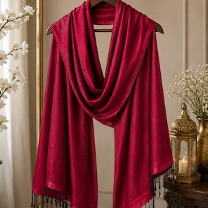 May include: A deep red scarf with a subtle tonal jacquard pattern, featuring a smooth border and black fringe tassels at both ends. The scarf is draped over a dark wooden hanger with the text PALISHQ COLLECTIONS printed in white.