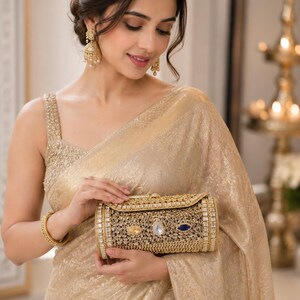 May include: A gold-toned cylindrical clutch bag featuring an intricate filigree metal overlay, embellished with clear, yellow, and blue faceted stones. The structured accessory has a metallic finish and a secure clasp closure on the top edge.
