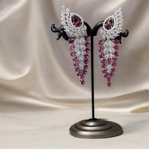 May include: A pair of dangle earrings with a pink and white crystal design. The earrings are on a stand.