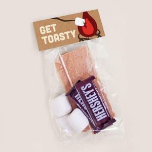 May include: A clear plastic bag containing the ingredients for a s'more. The bag includes a graham cracker, a Hershey's chocolate bar, and three marshmallows. A brown paper tag with the text "GET TOASTY" is attached to the bag.