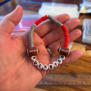 May include: A beaded bracelet featuring alternating red and gray flat circular clay beads, two brown football-shaped beads with white laces, and white round beads with black letters spelling out the word BUCKEYES in a curved arrangement.