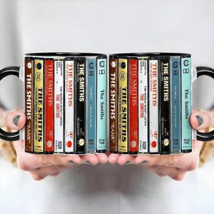 Puede incluir: Dos tazas de cerámica con asas y bordes negros, cada una con un diseño de cintas de cassette. Las cintas están etiquetadas con el nombre de la banda "The Smiths" y títulos de álbumes en varios colores. Un regalo único para los amantes de la música.