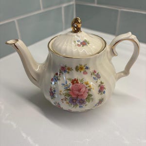 May include: Cream porcelain teapot with a floral pattern, including a pink rose and other colourful flowers. It has a gold handle, spout, and trim. The lid is topped with a gold finial. The teapot is a classic design.