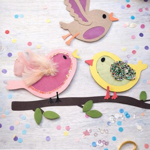 Può includere: Tre decorazioni di uccelli di carta colorata su una superficie di legno bianco, con coriandoli e forbici. Gli uccelli sono marroni, rosa e gialli, con dettagli come piume e glitter. Il testo in basso recita "Oster Basteldatei für Vögelchen direkt als Download."