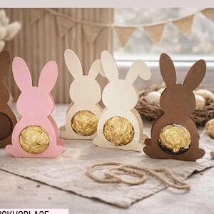Peut inclure: Découpes de lapins de Pâques en papier marron, rose et crème, chacune contenant une sphère de chocolat dorée. Les découpes sont disposées sur une surface neutre, avec des éléments décoratifs comme des œufs et de la ficelle.