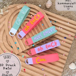 Könnte beinhalten: Fünf Silikon-Schlüsselanhänger in verschiedenen Farben wie Mint, Pink, Lavendel und Koralle. Jeder hat erhabenen Text: Oh, hi, oh fuCk, MORE AMORE, oh fuCk und Ciao. Die Schlüsselanhänger haben an einem Ende eine Schlaufe zur Befestigung.