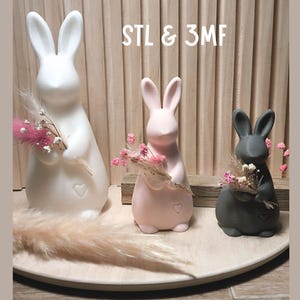 Puede incluir: Tres figuras de conejos de cerámica en blanco, rosa y gris, cada una sosteniendo un pequeño ramo de flores rosas. Los conejos tienen detalles en forma de corazón y están colocados sobre una bandeja de madera. El texto "STL & 3MF" es visible.