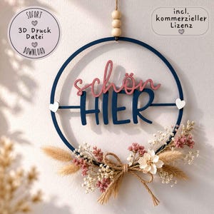 Könnte beinhalten: Ein kreisförmiger, dunkelblauer Wandbehang mit dem Schriftzug "schön HIER" in rosa und dunkelblauer Schreibschrift. Der Ring ist mit Trockenblumen, Gräsern und einer Juteschleife verziert. Oben hängen drei Holzperlen und zwei kleine weiße Herzen.