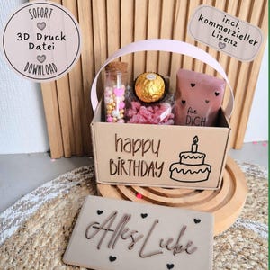Könnte beinhalten: Ein heller, rechteckiger Geschenkkorb mit Henkel, verziert mit dem Schriftzug 'happy BIRTHDAY' und einer Tortenabbildung. Darin befinden sich ein Glasröhrchen mit Perlen, eine goldene Praline und ein rosa Beutel mit der Aufschrift 'für DICH'. Ein passender Deckel trägt den Text 'Alles Liebe'.
