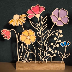 Op de afbeelding: Een houten wanddecoratie met een boeket kleurrijke bloemen met een houten basis. De bloemen zijn uit hout gesneden en geschilderd in tinten roze, geel, blauw en paars.