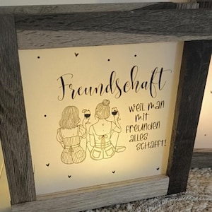 Könnte beinhalten: Ein beleuchtetes Schild in einem Holzrahmen mit weißem Hintergrund und einer schwarzen Illustration von zwei Frauen, die mit Weingläsern anstoßen. Der Text lautet "Freundschaft weil man mit Freunden alles schafft!"