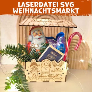 May include: A wooden Christmas market display box with a Santa Claus figure, candy cane, and other festive items. The box has a decorative roof and the words "Weihnachtsmarkt to go" and "Zauuberhafte Weihnachtszeit" are visible.