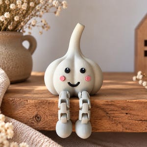 May include: A light-coloured garlic bulb figurine with a smooth surface, featuring a printed face with black eyes, a small black smile, and pink circular cheeks. The garlic has two articulated, segmented legs hanging down from the base.