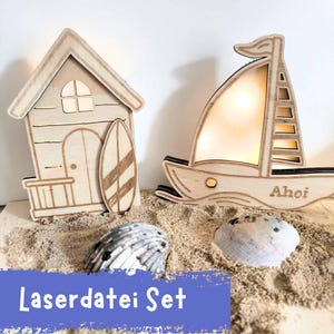 Puede incluir: Juego decorativo de madera con temática de playa que presenta una pequeña casa con una tabla de surf y un velero con la palabra "Ahoi". El juego incluye dos conchas marinas y se muestra sobre arena. El texto "Laserdatei Set" es visible.