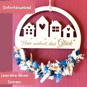 Könnte beinhalten: Ein halbrunder Holz-Türhänger mit drei ausgeschnittenen Häusern. Die Häuser haben Herzen darauf und der Text "Hier wohnt das Glück" ist darunter geschrieben. Der Hänger ist mit einem Kranz aus getrockneten Blumen in Blau- und Weißtönen verziert.