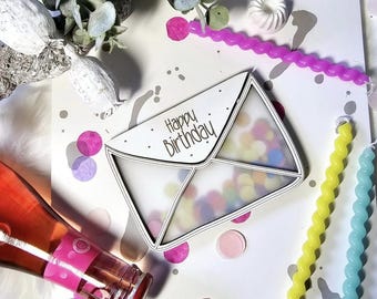 Może przedstawiać: Uroczysta kompozycja urodzinowa z białą kartką w kształcie koperty z napisem "Happy Birthday" i konfetti. Obraz zawiera butelkę różowego wina, spiralne świece i elementy dekoracyjne.