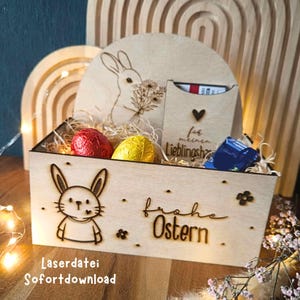 Può includere: Cesto pasquale in legno con disegni di coniglietti e fiori tagliati al laser. Il cesto contiene uova di cioccolato confezionate e un piccolo regalo. Le parole "Frohe Ostern" sono incise sul davanti. Testo "Laserdatei Sofortdownload".