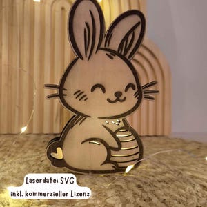 Puede incluir: Un conejo de madera cortado con láser sosteniendo un huevo. El conejo tiene una cara sonriente y está sentado sobre una superficie de madera. El texto "LaserdDatei SVG inkl. kommerzieller Lizenz" es visible en la parte inferior de la imagen.
