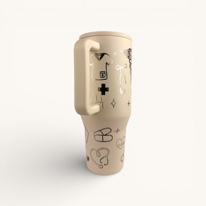 May include: A beige insulated tumbler with a handle, adorned with silver medical-themed illustrations. The design includes a nurse's uniform, a cross, a stethoscope, and a heartbeat line. The tumbler is designed to keep beverages cold or hot.