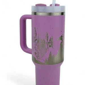 Puede incluir: Un vaso t&eacute;rmico lila con asa, tapa blanca y pajita. El vaso presenta un gr&aacute;fico plateado de un castillo, &aacute;rboles y una figura. El asa y la tapa son del mismo color lila que el cuerpo.