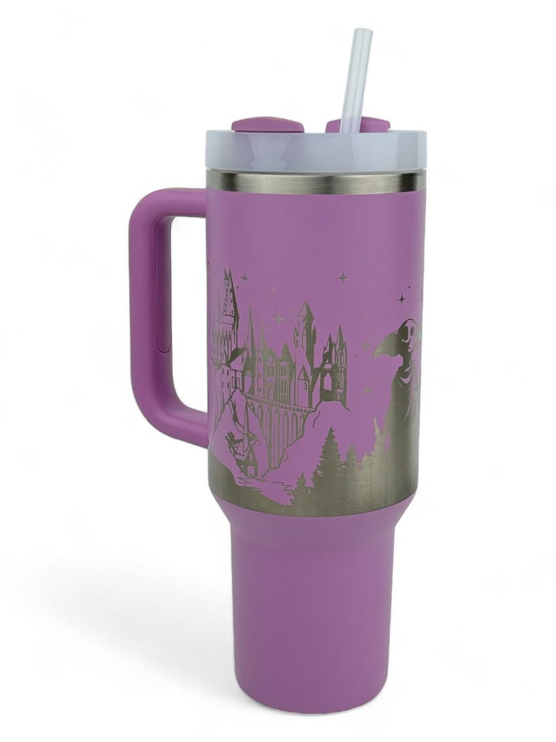 Puede incluir: Un vaso t&eacute;rmico lila con asa, tapa blanca y pajita. El vaso presenta un gr&aacute;fico plateado de un castillo, &aacute;rboles y una figura. El asa y la tapa son del mismo color lila que el cuerpo.