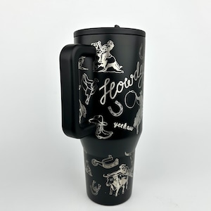 Puede incluir: Taza de viaje negra con asa y tapa, con ilustraciones plateadas con tem&aacute;tica de vaquero. El dise&ntilde;o incluye un vaquero montando a caballo, una herradura, una bota de vaquero y la palabra "Howdy" en cursiva.