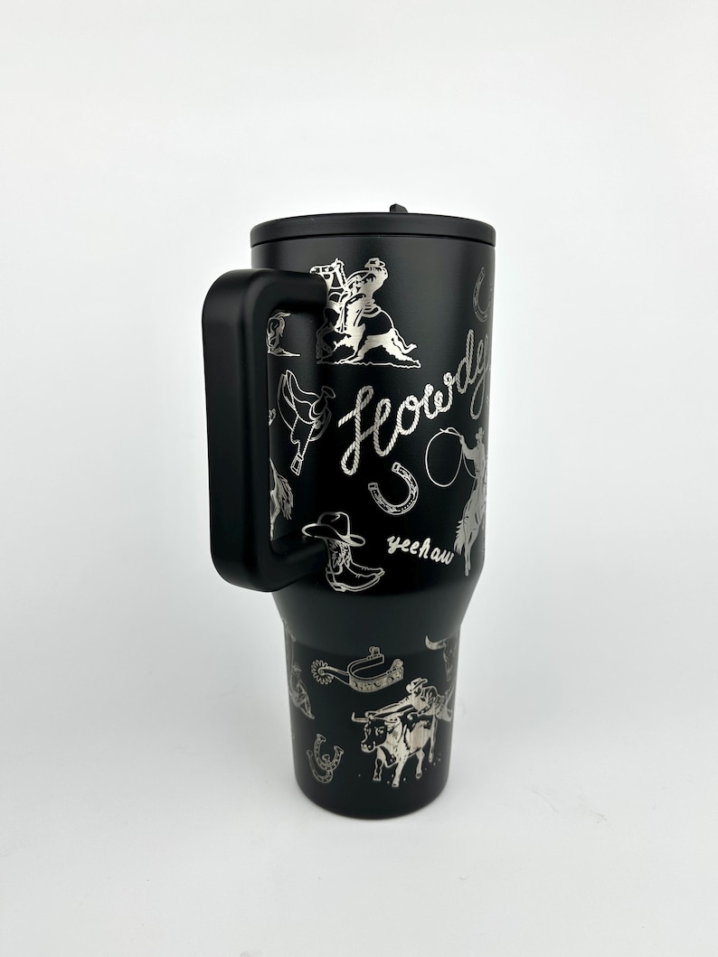 Puede incluir: Taza de viaje negra con asa y tapa, con ilustraciones plateadas con tem&aacute;tica de vaquero. El dise&ntilde;o incluye un vaquero montando a caballo, una herradura, una bota de vaquero y la palabra "Howdy" en cursiva.
