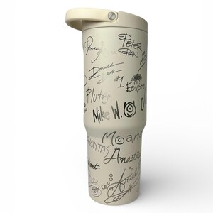 Puede incluir: Un vaso t&eacute;rmico de color crema con asa y tapa de rosca. El vaso est&aacute; cubierto de firmas y nombres en negro, incluyendo "Peter Pan", "Donald Duck" y "Ariel". El vaso est&aacute; dise&ntilde;ado para mantener las bebidas fr&iacute;as o calientes.
