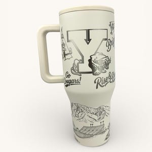 以下が含まれることがあります： クリーム色のハンドルと蓋付きのトラベルマグ。マグには、山脈、ティピー、クーガー、文字「Y」の白黒イラストが描かれています。「Go Cougars!」と「Rise & Shine」の文字が含まれています。