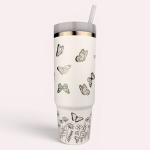 以下が含まれることがあります： グレーの蓋とストローが付いた白いタンブラー。タンブラーには、銀色の蝶と花の模様が施されています。冷たい飲み物用に設計されています。