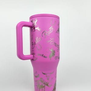 Puede incluir: Un vaso t&eacute;rmico rosa con asa, con gr&aacute;ficos plateados de tem&aacute;tica vaquera y texto, incluyendo el nombre "Bella Ruth" y la palabra "Yeehaw". El vaso est&aacute; dise&ntilde;ado para mantener las bebidas fr&iacute;as o calientes.